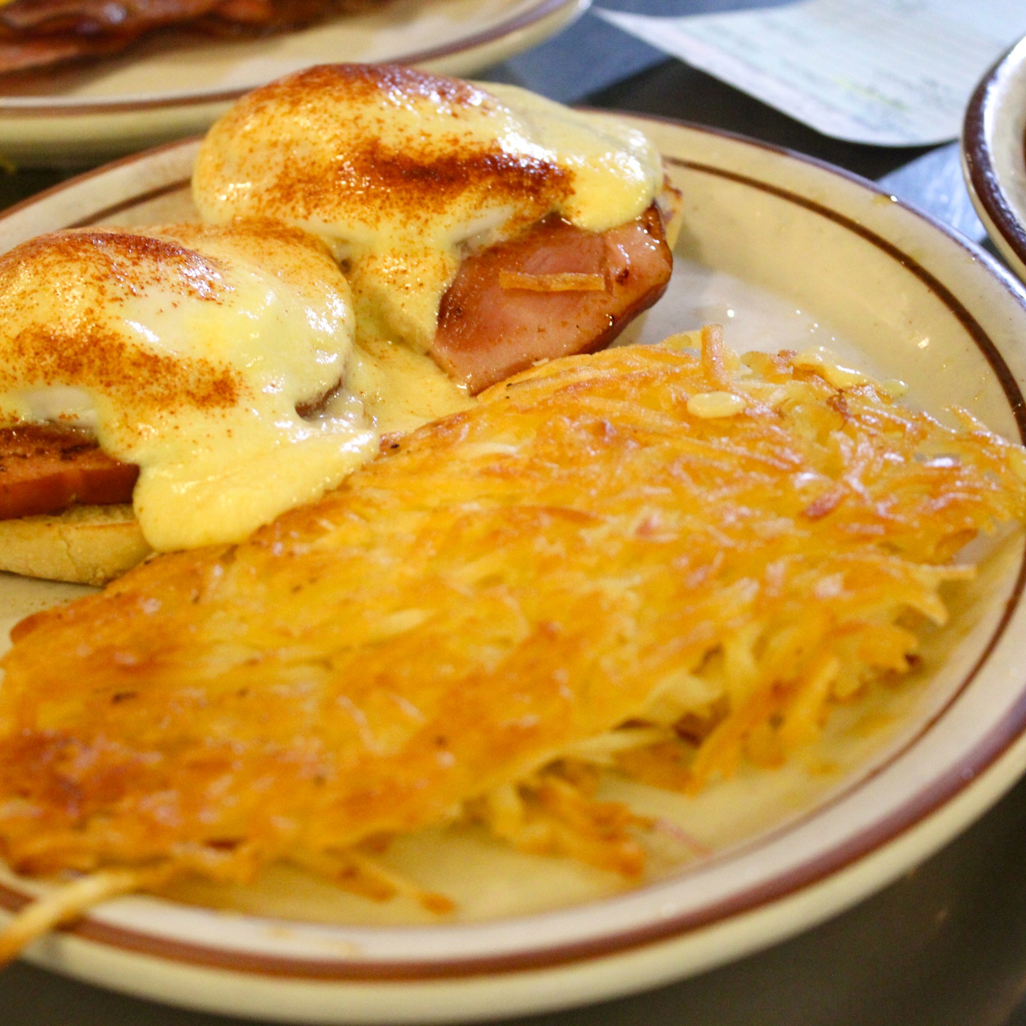 a plate of hashbrowns next to eggs benedict. Hollandaise topping two poached eggs and a slice of ham on a grilled english muffin